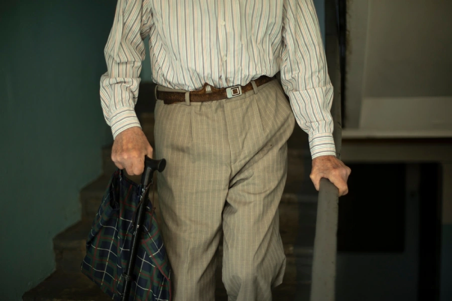 elder man walking down the stairs holding the railing in one hand and a grocery trolley in the other